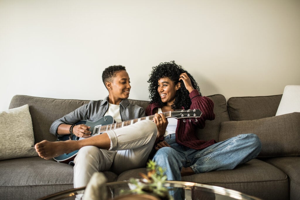 Lesbian couple do date night at home relaxing on couch with guitar