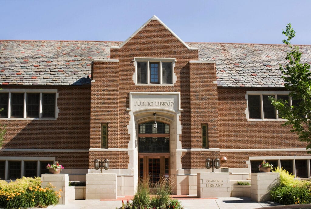 Front entrance of a small brick public library, with a copper and glass door and slate roof under a clear blue sky.